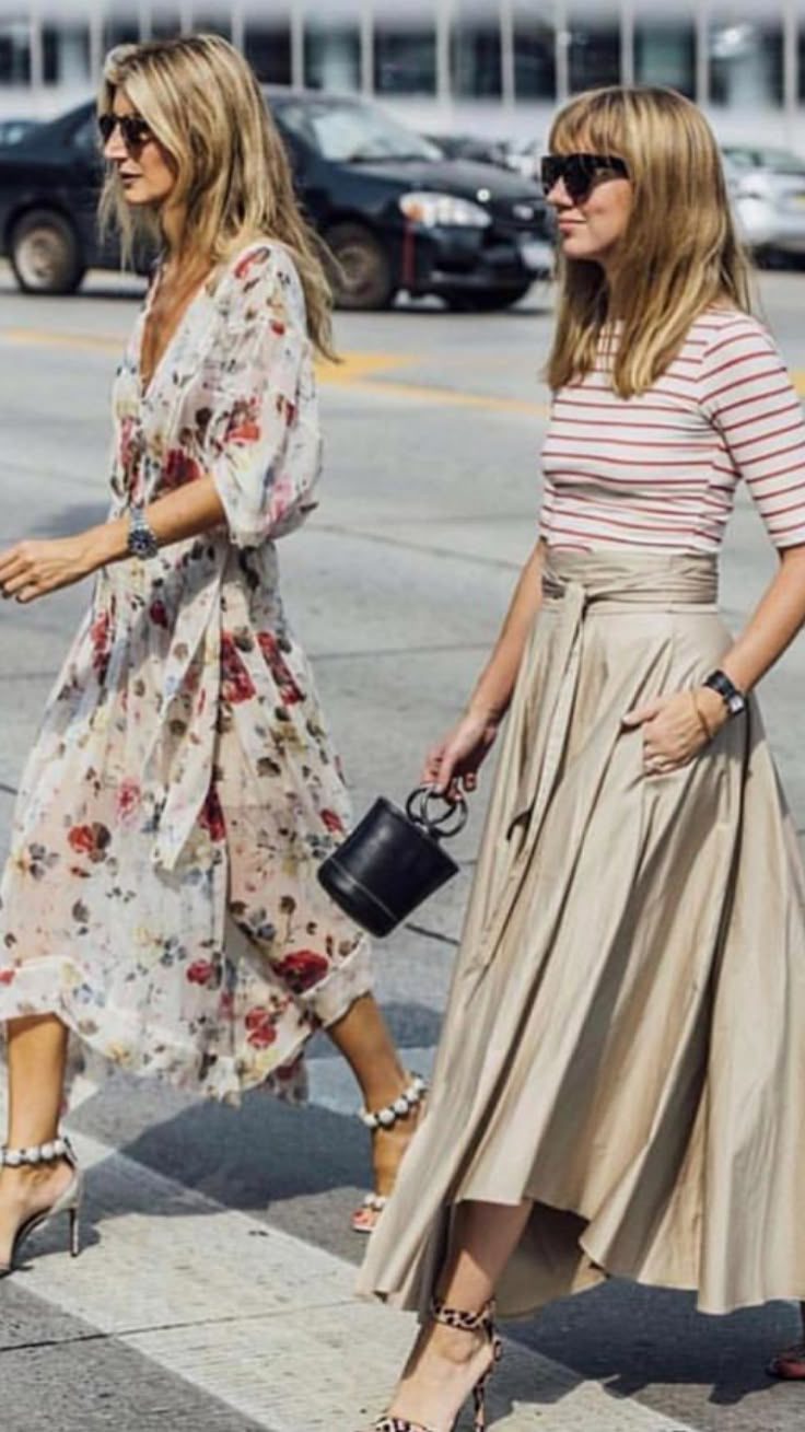 Two women in stylish outfits walk on a city street. One wears a floral dress, the other a striped top and beige skirt. They appear relaxed.