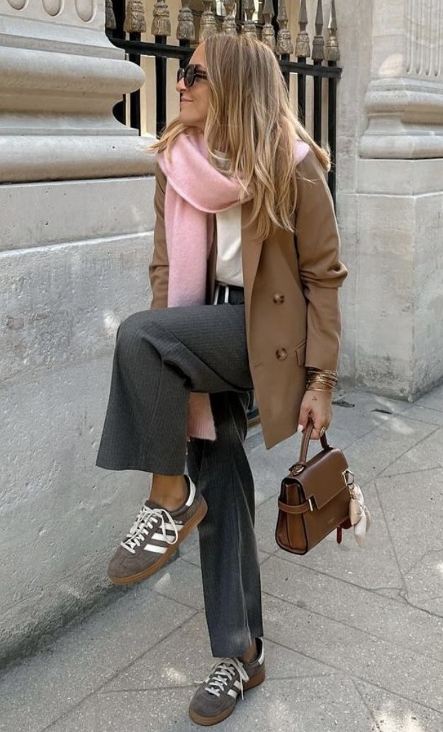 Woman in a brown coat, pink scarf, and grey pants leans against a stone wall, holding a brown handbag. Wearing sunglasses and sneakers.