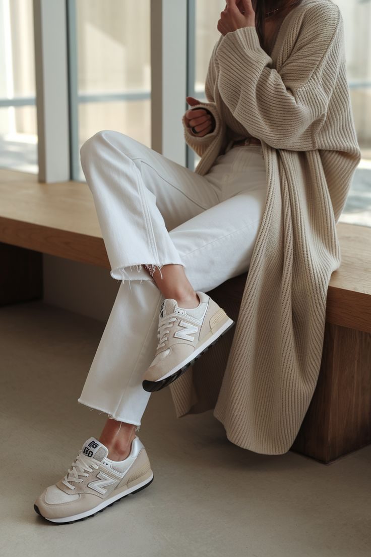 Woman in beige cardigan and white pants sits on wooden bench, wearing beige sneakers with "N" logo. Sunlit room, relaxed mood.