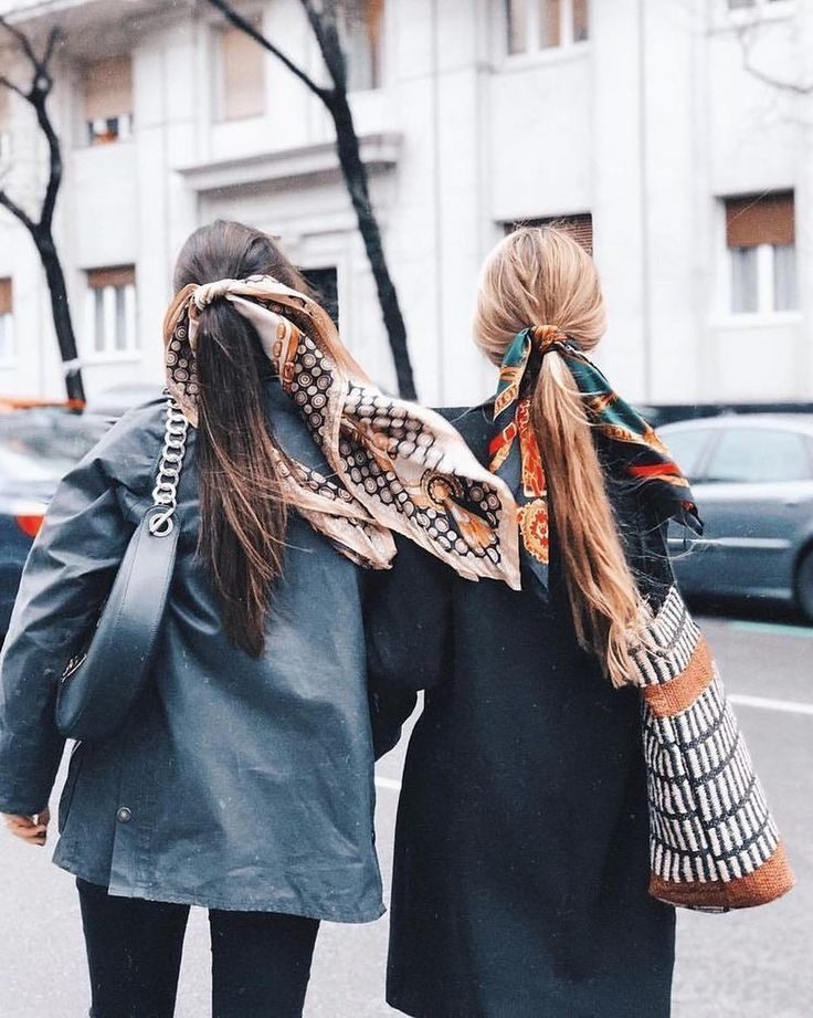 Two women with scarves in their hair walk arm in arm on a city street. One holds a black bag; the background shows a building and cars.