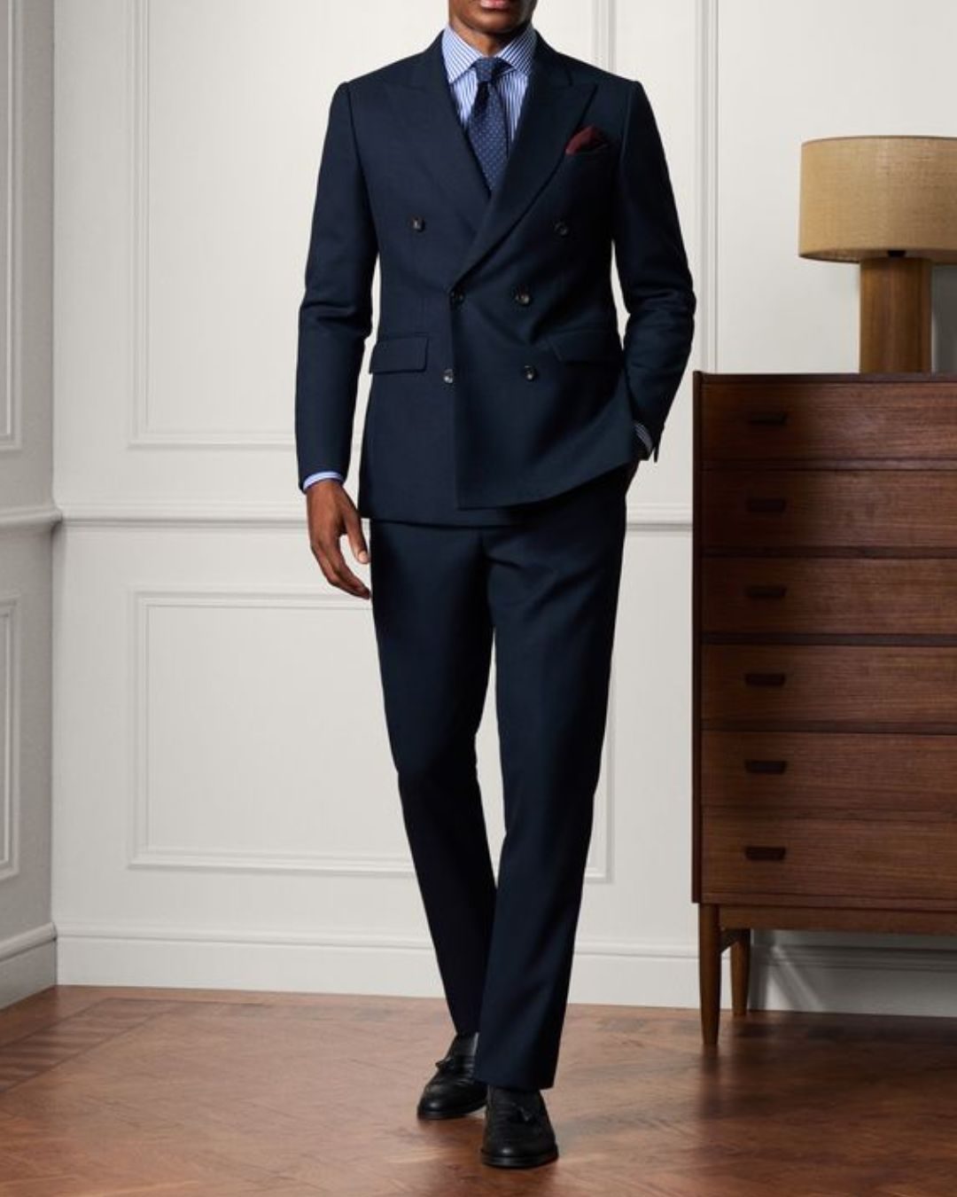 Man in a dark double-breasted suit and tie stands confidently in a room with a wood dresser and lamp. Neutral tones set a formal mood. Attending a black tie formal wedding event.