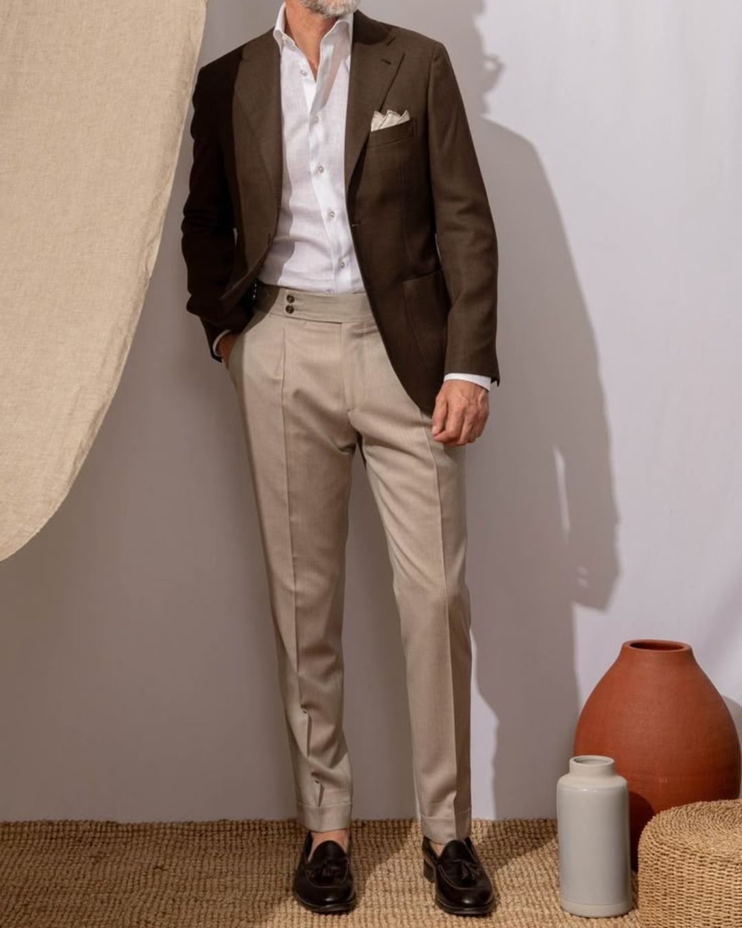 Man in brown blazer and beige pants stands on woven mat. Attending a garden party wedding.
