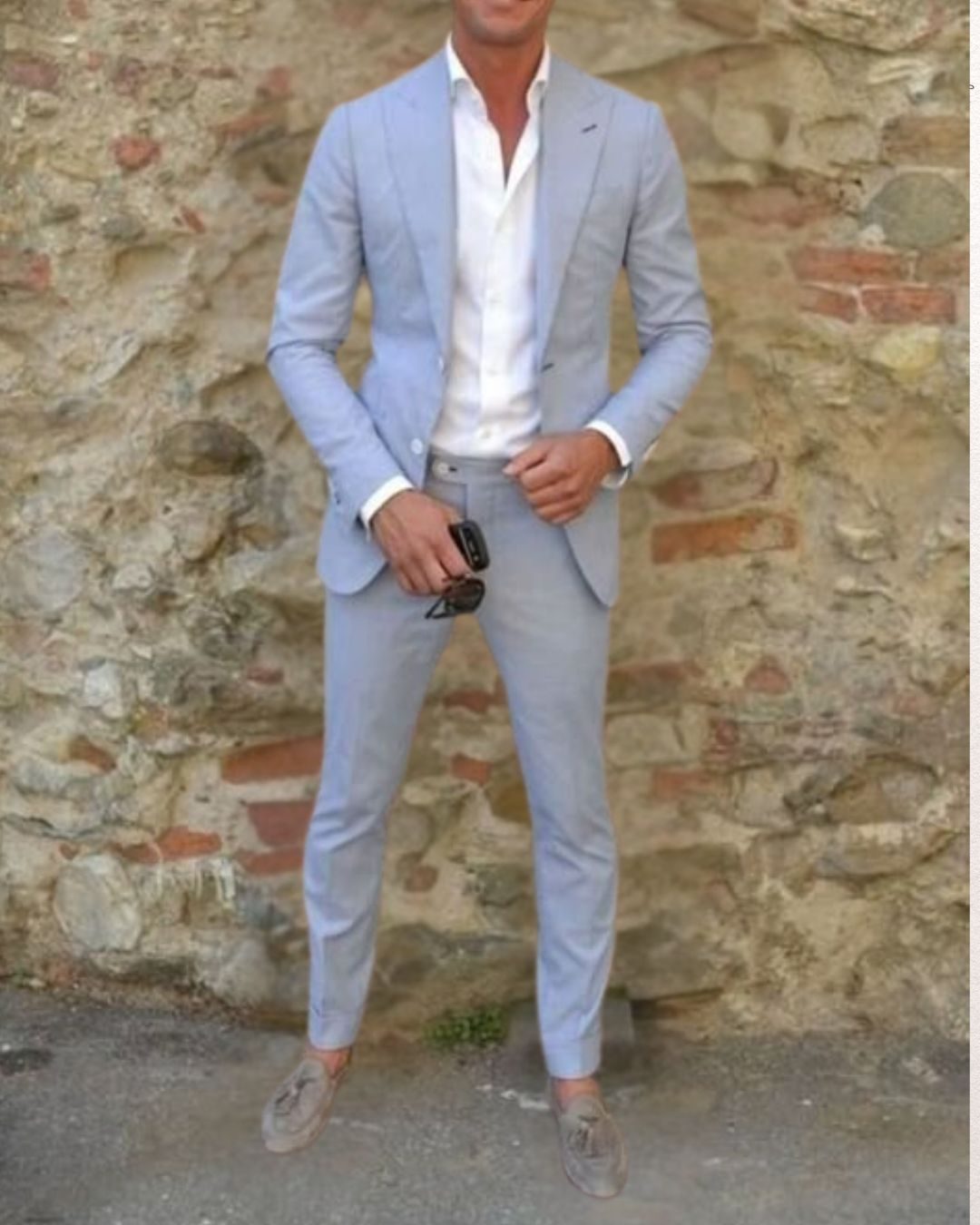 Man in light blue suit and white shirt holds sunglasses, standing against a rustic stone wall. He's wearing grey loafers, exuding a relaxed vibe. He is attending a beach wedding.